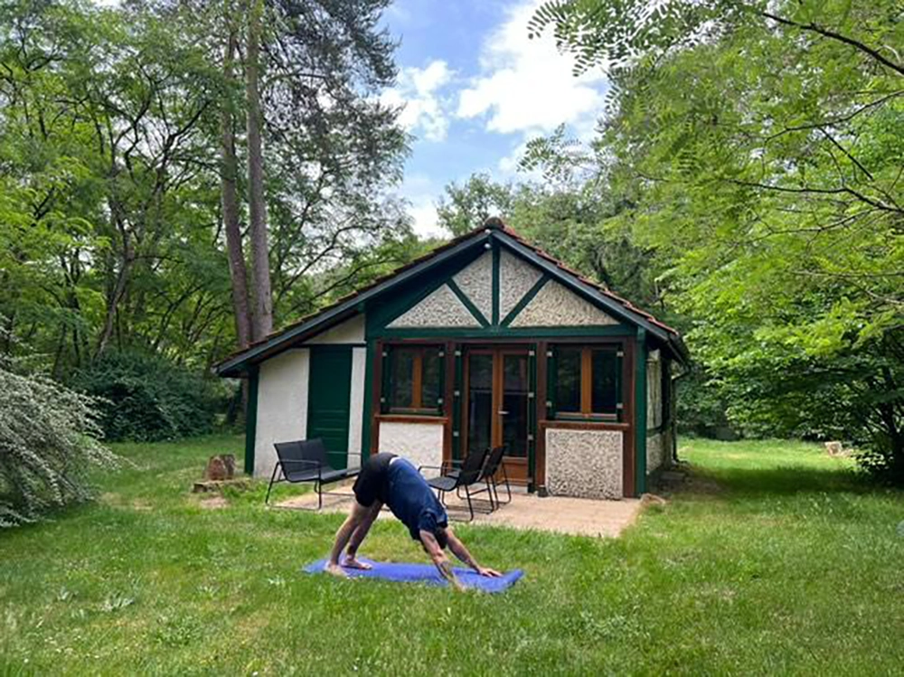 Outdoor view of the apartment in the background with on the foreground a white male in his thirties wearing a blue t-shirt and a black short doing the down dog yoga pose on a blue mat on the grass.