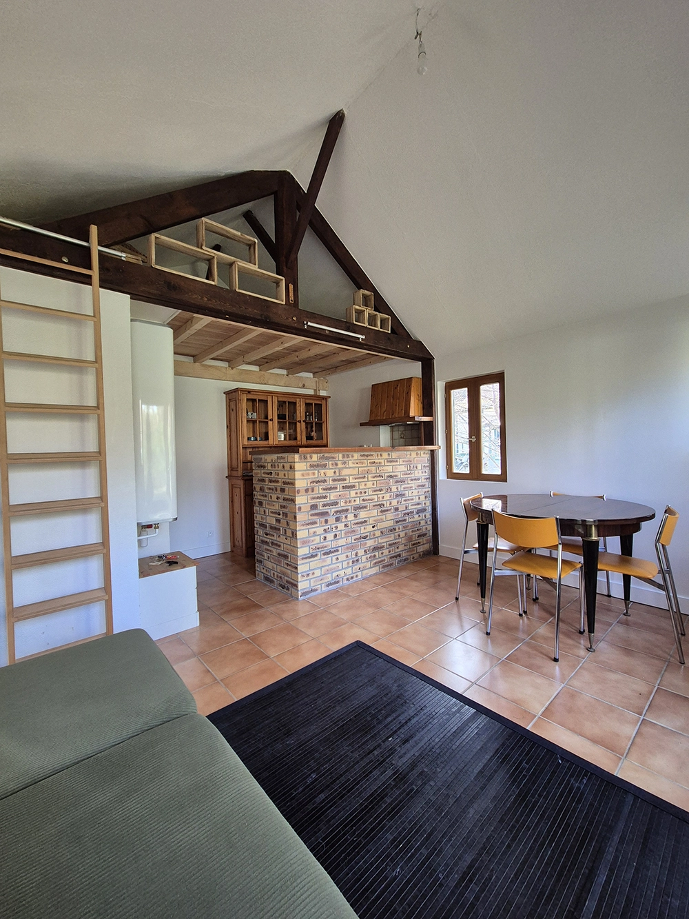 Indoor view of the apartment, tree seats light green and yellow sofa on the left, black carpet and a brown table with four yellow chairs on the right. Kitchen behind the table, above the kitchen there is a ladder to access the mezzanine.